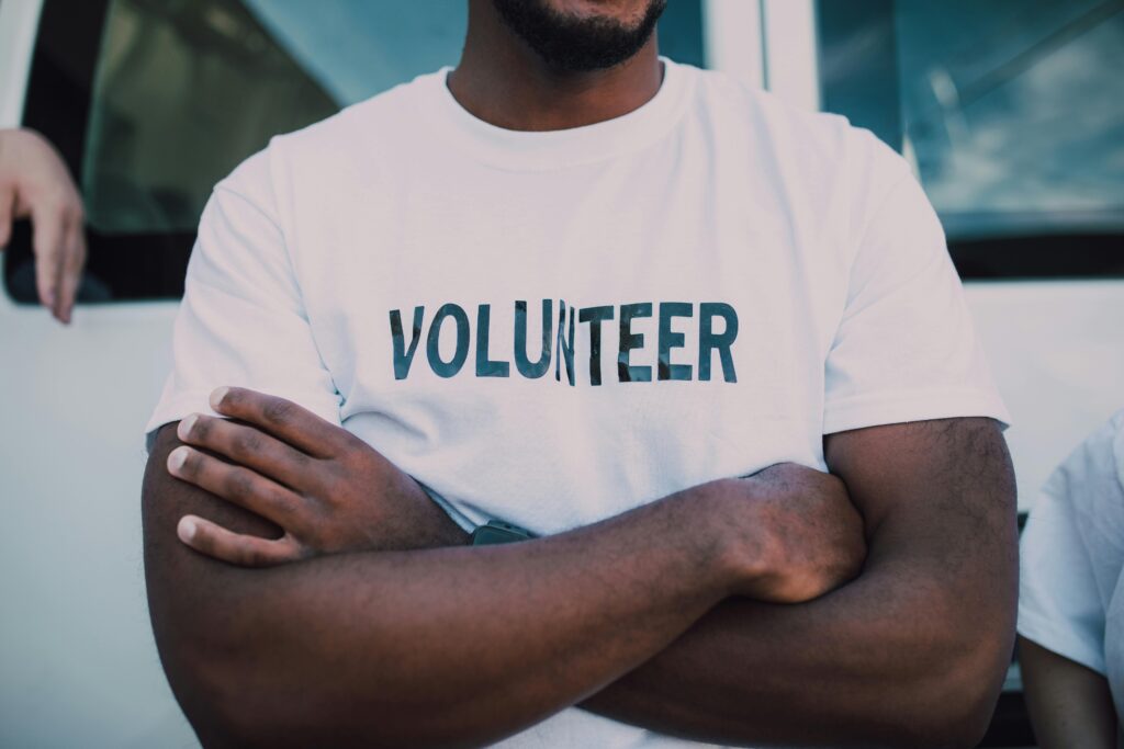 person with dark skin wearing a white short with the word "volunteer" in all caps on the front, crossing their arms across their chest. Photo by RDNE Stock project.